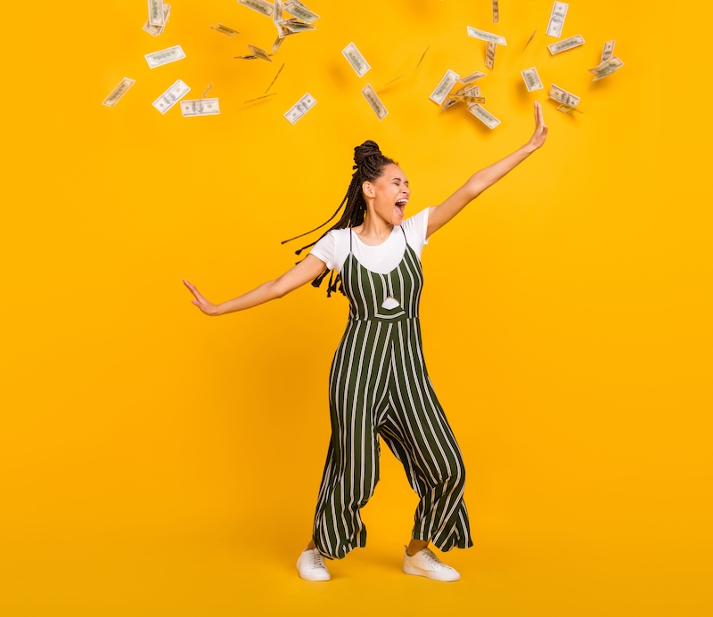 Glückliche junge Frau tanzt im Geldregen vor gelbem Hintergrund.